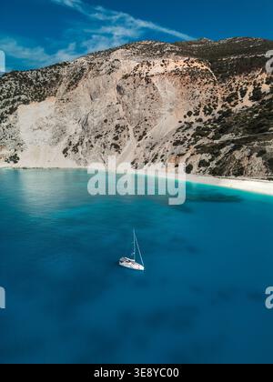Blick von der Drohne auf Myrtos Beach, Kefalonia, Griechenland, mit einem einsamen Segelboot auf lebhaftem türkisfarbenem Wasser und dramatischen Kalksteinbergen im Oktober. Stockfoto