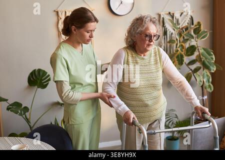 Seniorin mit Gehhilfe, junge erwachsene Betreuerin, die Mobilität während der Rehabilitation von Pflegeheimen unterstützt Stockfoto