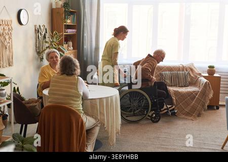 Seniorenmann mit Behinderung, der aus dem Rollstuhl wechselt, mit Betreuungspersonal im Pflegeheim, während Seniorinnen am Tisch sprechen Stockfoto
