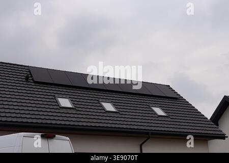 Eine Reihe von Photovoltaik-Solarpaneelen auf einem schwarz gekachelten Dach mit drei Dachfenstern. Ein weißer Lieferwagen ist teilweise im Vorfeld sichtbar Stockfoto