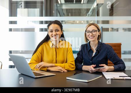 Zwei lächelnde, multiethnische Geschäftsfrauen sitzen mit Laptop, Tablet und Dokumenten an einem Konferenztisch und repräsentieren Teamarbeit, Technologie, und Erfolg in einer modernen Büroumgebung Stockfoto