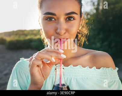Junge Frau in blauen Gläsern, die durch einen Strohhalm trinkt und in die Kamera blickt, mit warmem Sonnenuntergangslicht im Hintergrund. Stockfoto