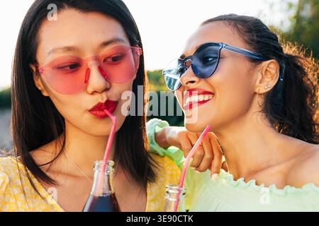 Weibliche Freunde mit Getränken. Nahporträt von zwei Frauen, die im Freien Brillen in verschiedenen Farben tragen. Stockfoto