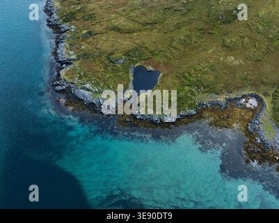 Aus der Vogelperspektive auf die zerklüftete Küste der Lofoten mit felsigen Klippen, türkisfarbenem Wasser und einem kleinen herzförmigen Teich in der grünen Tundra Nordland, Norwegen. Stockfoto
