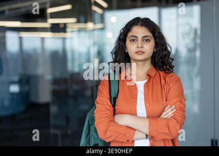 Junge Frau mit ernstem Ausdruck, mit orangefarbenem Hemd und Rucksack, die Entschlossenheit und Ehrgeiz in einer akademischen oder beruflichen Tätigkeit vermittelt Stockfoto