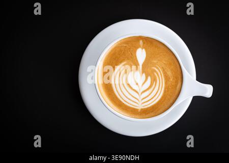 Frisch gebrühter Latte Coffee mit cremiger Schaumkunst in einer weißen Tasse, serviert in einer raffinierten dunklen Umgebung. Stockfoto