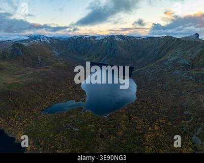 Aus der Vogelperspektive auf die zerklüftete Landschaft von Andoya mit einem dunklen Bergsee umgeben von steilen Gipfeln und schneebedeckten Graten unter einem dramatischen Himmel Stockfoto