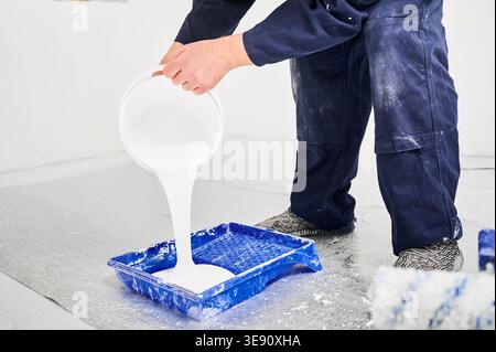 Arbeiter gießen weiße Beschichtung in blaue Schale auf grundierter Bodenoberfläche, Nahaufnahme. Vorbereitung der Lackierung für die Endbearbeitung, den Arbeitsfluss für die Innenraumsanierung und das Konzept der Oberflächenbehandlung Stockfoto