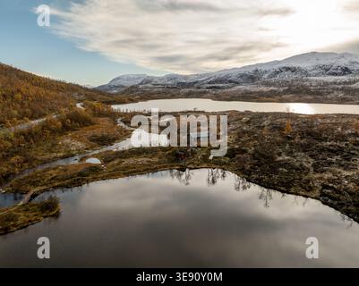 Aus der Vogelperspektive einer kleinen Holzhütte, eingebettet zwischen ruhigen Seen und herbstfarbenen Hügeln mit schneebedeckten Bergen unter einem bewölkten Himmel in Senja, Troms, Stockfoto