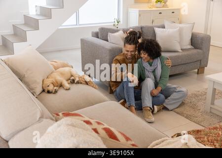 Pärchen in Pullis sitzen auf gefliestem Boden im Wohnzimmer, Mann zeigt Telefon während Hündchen schläft. Gemütlich, sonnenbeleuchtet, natürlich, Zuhause, Lounge, Hund, Retriever Stockfoto