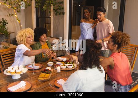 Verschiedene Freunde, die zwanglos gekleidet sind, ziehen Gläser auf und gießen Wein auf der Veranda mit Cupcakes. Im Freien, Terrasse, Treffen, Wurstwaren, Dessert, Be Stockfoto