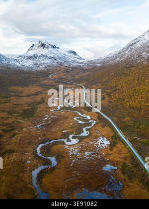 Aus der Vogelperspektive auf einen sich windenden Fluss und eine lange Straße, die sich durch ein Tal mit schneebedeckten Bergen und Herbstlaub in der Landschaft von Senja, TR. Erstreckt Stockfoto