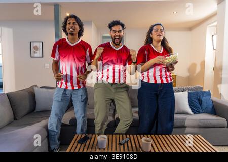 Verschiedene Freunde tragen passende rote weiße Trikots, jubeln, halten Popcorn zu Hause auf dem grauen Sofa. Trio, Teamwear, Watchparty, fotorealistisch, Sport Stockfoto
