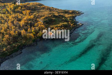 Blick aus der Vogelperspektive auf Hamn i Senja mit einem roten Bootshaus am felsigen Ufer, umgeben von goldenen Herbstbäumen und türkisfarbenem Wasser in Skaland, Troms, Norw Stockfoto