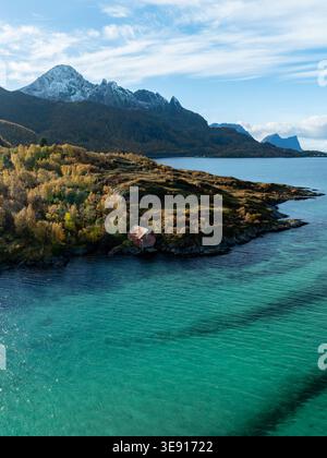 Aus der Vogelperspektive eines roten Bootshauses am felsigen Ufer von Hamn i Senja, umgeben von Herbstbäumen und türkisfarbenem Wasser mit schneebedeckten Bergen in Skaland Stockfoto