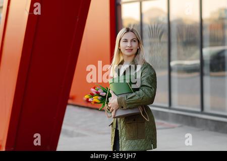 Junge blonde Frau mit einem Blumenstrauß aus bunten Tulpen und einer grünen Hutschachtel, die durch die Stadt in der Nähe einer roten modernen Architektur spaziert Stockfoto