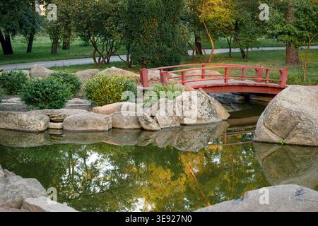 Eine ruhige japanische Gartenszene mit einer kleinen roten Brücke über einen Teich, umgeben von großen beigen Felsen, lebhaften grünen Büschen und Bäumen reflektieren Stockfoto