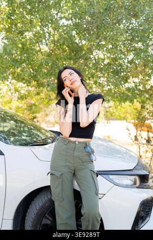 Besorgte junge Frau, die neben ihrem kaputten weißen Auto in einem ländlichen Waldgebiet stand und auf ihrem Handy Pannenhilfe anrief. Stress und Co Stockfoto