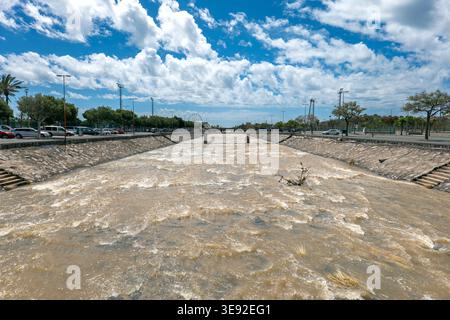 Regenwasserkanal auf Gran Canaria, der nach starkem Regen schnell fließendes, schlammiges Wasser mit einer kleinen Brücke und blauem Himmel im Hintergrund transportiert. Stockfoto