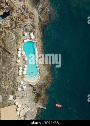 Blick aus der Vogelperspektive auf ein Resort-Schwimmbad auf einer felsigen Klippe mit weißen Sonnenschirmen und türkisfarbenem Wasser neben dem Mittelmeer in Taormina, Sizilien, Stockfoto