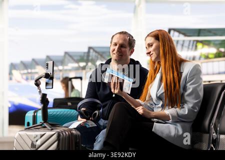 Mann und Frau Influencer erstellen Reisevlog am Flughafen, halten Telefon und nehmen Video auf, während sie sich auf den Abflug vorbereiten. Präsenz in sozialen Medien, die ihre Online-Reise dokumentieren. Stockfoto