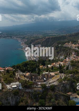 Aus der Vogelperspektive des antiken Theaters der Ruinen von Taormina auf einer Klippe mit Blick auf die Mittelmeerküste und die Stadt unter einem bewölkten Himmel Taormina, Sici Stockfoto