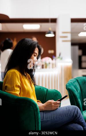 Frau, die im eleganten Hotelfoyer sitzt und ein mobiles Gerät benutzt, während sie darauf wartet, in das moderne Resort einzuchecken. Asiatische Besucher mittleren Alters bereiteten sich bereit, ein Zimmer in einem modernen Loungebereich zu buchen Stockfoto