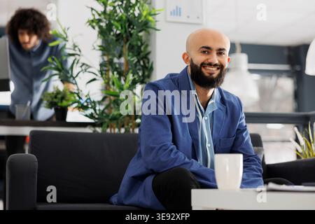 Lächelnder arabischer Geschäftsunternehmer, der in die Kamera schaut, während er im Büro auf der Couch sitzt. Start-up-Unternehmen erfolgreicher Manager mit Anzug Porträt im Coworking Space Stockfoto