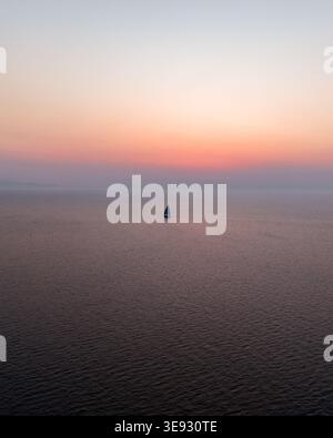Aus der Vogelperspektive eines einsamen Segelbootes, das auf dem ruhigen Mittelmeer unter einem trüben orangefarbenen Sonnenuntergangshimmel in Isola delle Femmine, Sizilien, Italien, segelt. Stockfoto