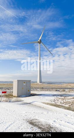 Luftaufnahme einer großen Windturbine und eines Versorgungsgebäudes in einer verschneiten Küstenlandschaft unter einem blauen Himmel mit hellen Wolken Urk, Flevoland, ne Stockfoto