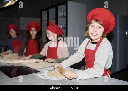 Porträt von Kindern, die Kochmütze und Schürzen tragen und Teig während des Kochkurses in der Küche Rollen. Unterstützung von kulinarischer Bildung, Teamarbeit, Essenswerkstatt, Elternschaft oder Schulförderung Stockfoto
