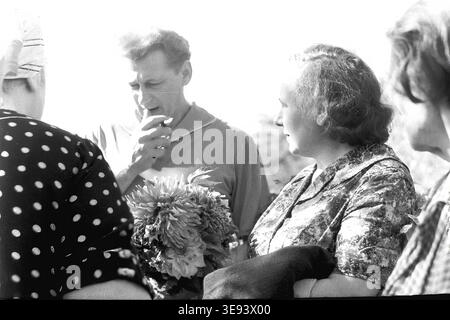 Historisches Schwarzweißfoto aus dem Archiv, das eine Nahaufnahme der Frauen in gemusterten Kleidern und eines Mannes, der während der feierlichen Eröffnungszeremonie des Denkmals für die Partisanenbewegung am Massengrab (späteres Karnauchow-Denkmal) in der Kalinina-Straße (heute Uchitelskaja), der Stadt Slawjansk, 1950er, raucht Die Teilnehmer führen ein zwangloses Gespräch unter dem hellen Sonnenlicht und unterstreichen die authentische Atmosphäre der Nachkriegszeit. Das Leben vor dem Krieg, der friedliche Donbass. Stockfoto
