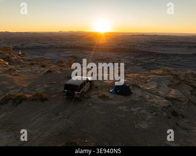 Aus der Vogelperspektive eines Geländefahrzeugs und eines Campingzelt auf einer felsigen Klippe mit Blick auf die riesigen Wüstenbadlands bei einem goldenen Sonnenaufgang in Hanksville, Uta Stockfoto