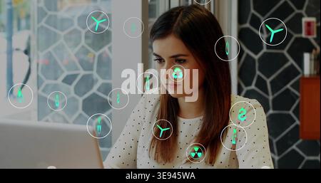 Arbeitende Frau in weißer Polka-Dot-Bluse, die am Arbeitsplatz auf einem Laptop schreibt, mit grünen Symbolen Stockfoto