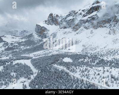 Aus der Vogelperspektive auf den schneebedeckten Sellapass mit gewundenen Bergstraßen, dichten Kiefernwäldern und zerklüfteten Gipfeln unter einem dramatisch bewölkten Himmel in den Dolomi Stockfoto