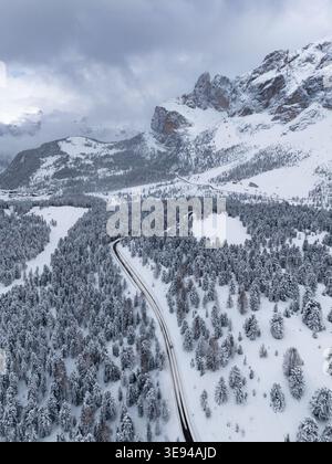Aus der Vogelperspektive auf den Sellapass, der sich durch einen schneebedeckten Kiefernwald mit zerklüfteten Berggipfeln unter bewölktem Himmel in Wolkenstein, Trentino-Südtirol, Italien schlängelt Stockfoto