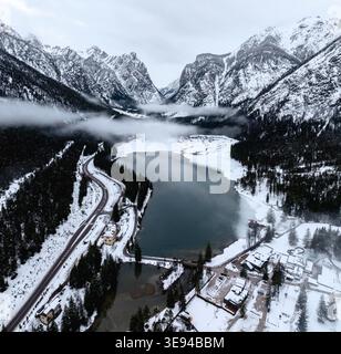 Aus der Vogelperspektive auf den Toblacher See, umgeben von schneebedeckten Bergen, dichten Kiefernwäldern und einer gewundenen Straße unter einem nebeligen Himmel in Toblach, Trentino-Süd Stockfoto
