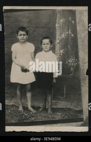 Schwarz-weißes Atelierporträt zweier barfuß russischer Kinder, die in der UdSSR in den 1930er Jahren vor gemalter Kulisse standen Der Junge und das Mädchen tragen einfach Stockfoto