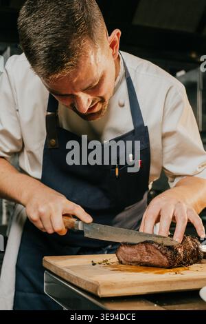 Ein Koch schneidet ein Stück Fleisch auf einem hölzernen Schneidebrett. Der Koch trägt eine blaue Schürze und ein weißes Hemd Stockfoto