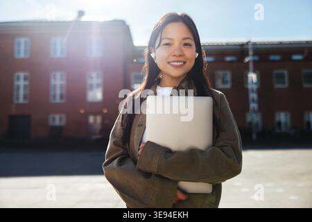 Ein lächelnder Student hält einen Laptop in der Nähe, während er auf einem sonnigen College-Campus schnurlose Ohrhörer trägt. Er vermittelt Selbstvertrauen, modernes Lernen und Studium im Freien Stockfoto