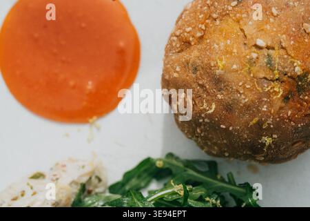 Frisch gebackenes Brot mit goldener Kruste und gestreuten Kräutern. Helle Orangensoßen-Pools neben dem Brotlaib. Knuspriger grüner Rucola und Weißkäse verleihen Farbe A Stockfoto