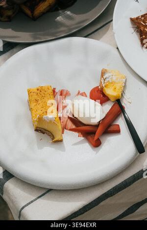 Delikates Dessert auf einem einfachen weißen Teller. Gelbe Kuchenscheibe mit kandiertem Topping. Rosafarbene Karottenstäbchen und Weichkäsekugel. Der schwarze Löffel liegt neben dem Stockfoto