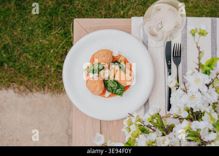 Die weiße Tafel fasst orangene Ravioli mit Grün und Creme. Ein Glas Wein steht neben Gabel und Messer. Blumen blühen in der Nähe eines Holztisches. Gras umrahmt den Stockfoto