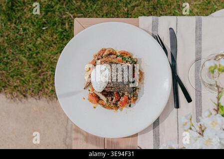 Frisch gebratener Fisch liegt auf einem farbenfrohen Gemüsebett. Der weiße Teller liegt auf einem Holztisch neben ordentlich platziertem Besteck. Gras und Blüte Stockfoto