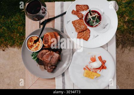 Gourmet-Mahlzeit auf einem rustikalen Holztisch. Reichhaltiges Steak, geröstetes Wurzelgemüse und frisches Gemüse. Ein Glas dunkler Rotwein ergänzt das fest. Plat Stockfoto