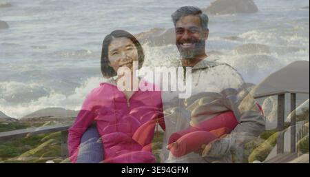 Ein stehendes Paar trägt rosa und graue Hoodies auf der Promenade und hält gerollte Yogamatten in der Nähe der Wellen Stockfoto