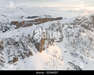 Aus der Vogelperspektive auf schneebedeckte zerklüftete Berggipfel und felsige Klippen unter einem nebeligen Himmel mit sanftem goldenem Licht in Wolkenstein in Gröden, Trentino-Alto Adi Stockfoto