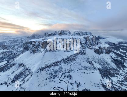 Aus der Vogelperspektive auf die schneebedeckte Sellagruppe mit einer gewundenen Straße und Wolken bei Sonnenuntergang in Wolkenstein in Gröden, Trentino-Südtirol, Ital Stockfoto