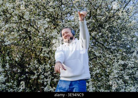 Fröhlicher bärtiger Mann, der im Frühling mit Kopfhörern in der Nähe eines blühenden Baumes tanzt Stockfoto