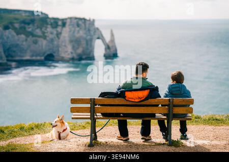 Dad und Sohn genießen die Zeit zusammen auf der Bank, Corgi-Hund ruht in Etretat Stockfoto